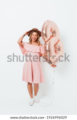 Happy international women's day, full length portrait of cheerful young woman wearing pink dress and straw hat holding golden number eight balloon on white studio background.