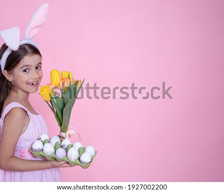 Little girl with easter bunny ears holds a bouquet of tulips and a tray of eggs in her hands on a pink studio background.