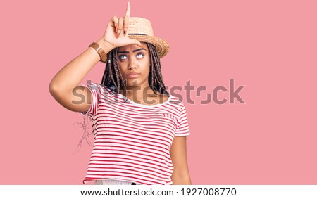 Young african american woman with braids wearing summer hat making fun of people with fingers on forehead doing loser gesture mocking and insulting. 