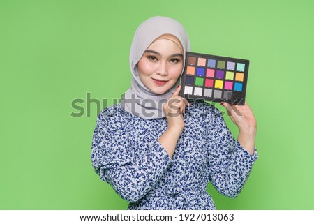 Young Asian girl holding color checker card, isolated on green background. Professional photography equipment concept.