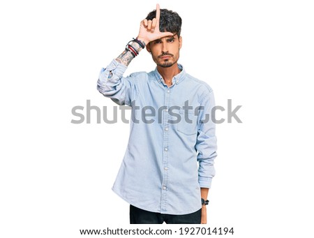 Young hispanic man wearing casual clothes making fun of people with fingers on forehead doing loser gesture mocking and insulting. 