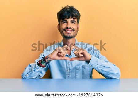 Young hispanic man wearing casual clothes sitting on the table smiling in love doing heart symbol shape with hands. romantic concept. 