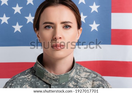 Portrait of soldier in camouflage looking at camera near american flag blurred at background 