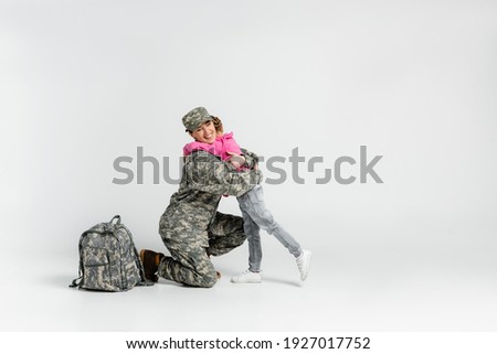 Cheerful soldier hugging daughter near backpack on grey background 