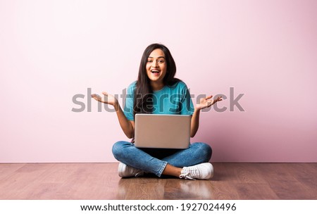 Indian asian young woman or girl sitting with laptop on her lap against pink wall on wooden floor Royalty-Free Stock Photo #1927024496
