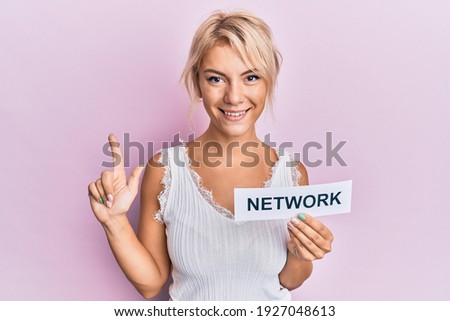 Young blonde girl holding paper with network word smiling with an idea or question pointing finger with happy face, number one 
