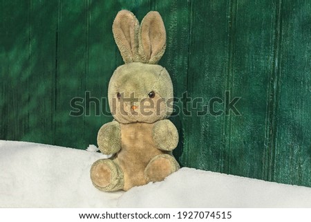 one old plush toy hare sits on a white snowdrift near a green wooden wall on a winter street