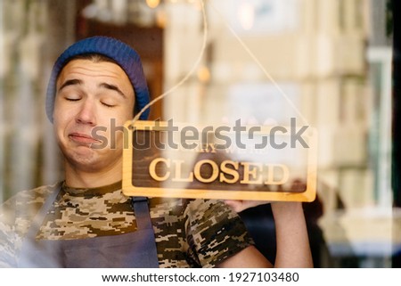Close up of young caucasian man wearing blue cap and grey apron About to close business in his own coffee shop and hold a sign to close his own shop. Funny face emotion.