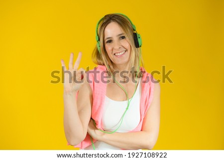 Young woman wearing headphones standing over yellow background smiling positive doing okay sign with hand and fingers. Successful expression.