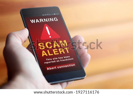 Closeup of male hand holding modern mobile phone with red screen and warning icon with the text "SCAM ALERT: This site has been reported as fraudulent. Abort connection". Concept of internet security. Royalty-Free Stock Photo #1927116176