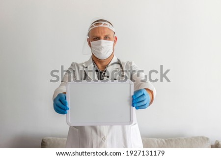 Young Caucasian doctor in white uniform holding whiteboard over grey background. Doctor showing medical sign whiteboard standing in half length. Male doctor with the blank board in the hospital.