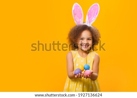 A wide smiling african girl in sunny orange dress with rabbit ears on her head with painted eggs in her hands. on a yellow background.