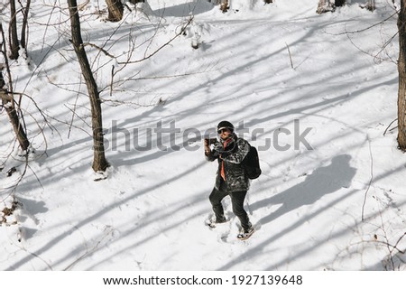 An adult man, a wanderer, a tourist in black warm clothes and a large backpack on his back travels through the snowy forest and takes pictures of the beautiful nature on a mobile phone, smartphone.