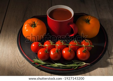 The dark food photography of tomatoes and red cup of tomato juice on the wooden kitchen table. Orange tomatoes and branch of cherry tomatoes around cup of tomato juice.