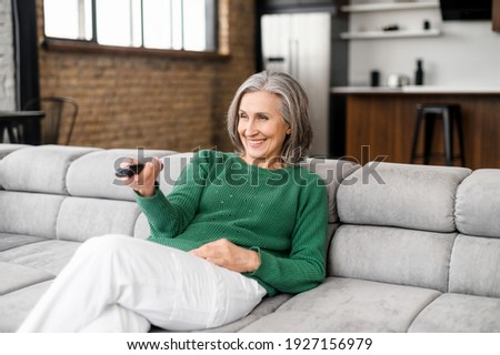 Happy smiling senior lady with gray hair in a green jumper sitting on the couch in the living room, pointing a tv remote controller to switch channels, excited to watch favorite movie online series