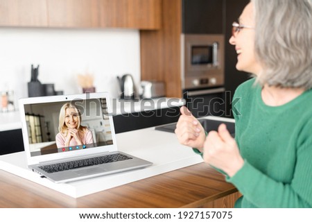 Senior mature middle-aged woman in glasses sitting in the kitchen, talking and looking at the screen with female friend image, showing okay sign, approving, staying at home and connected in pandemic
