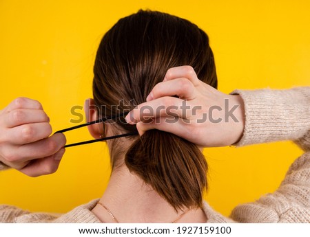 Back view shot of a fair-haired lady. There's a black rubber band on her hair with a black fake hair braid. The photo was taken against a yellow background. Royalty-Free Stock Photo #1927159100