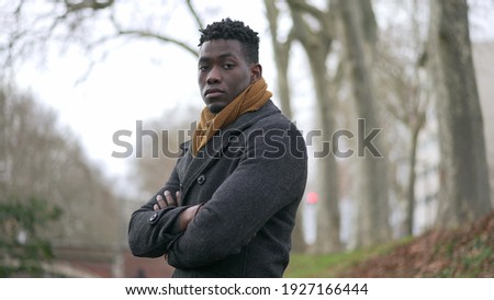 Black man arms crossed standing outside at park thinking wearing winter coat and scarf