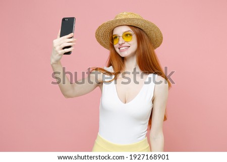 Young smiling nice redhead woman ginger long hair wearing straw hat glasses summer clothes doing selfie shot on mobile phone talking by video call isolated on pastel pink background studio portrait