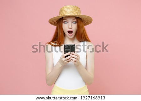 Young surprised caucasian pretty nice redhead woman 20s ginger long hair in straw hat summer clothes hold in hand using chatting by mobile cell phone isolated on pastel pink background studio portrait