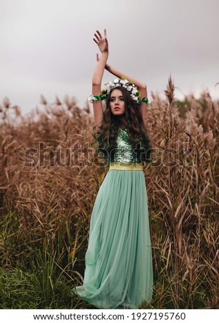The forest fairy with long dark hair and a wreath of flowers in a long turquoise dress near reeds.The forest nymph. Halloween costume.