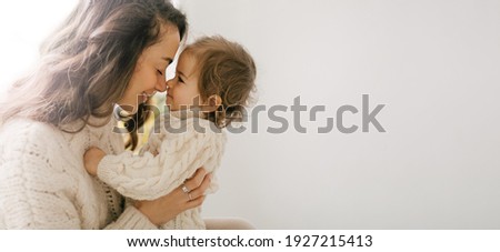 Young mother hugging her little daughter, they sitting near window and smiling. 