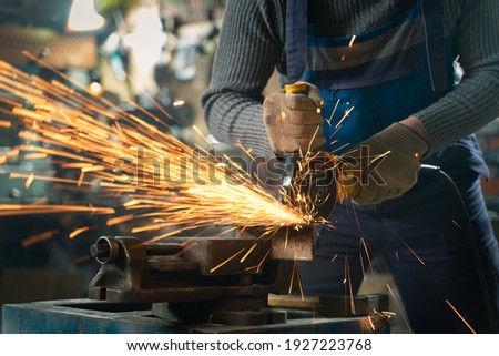 Locksmith in special clothes and goggles works in production. Metal processing with angle grinder. Sparks in metalworking. Royalty-Free Stock Photo #1927223768