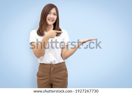 Beautiful asian woman standing wear white t-shirt holding something on palm and point away side looking to hand isolated on blue background with copy space and clipping paths.