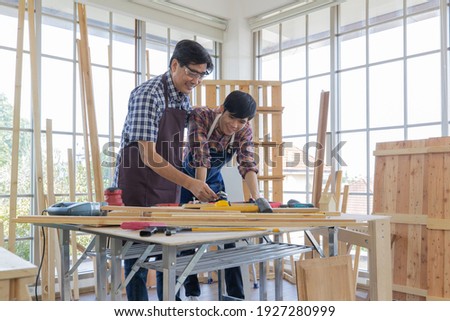 Two asian adult workers wearing plaid shirt stand smiling happily leaning over wood working desk together while assemble the wooden furniture with stainless wrench. Royalty-Free Stock Photo #1927280999