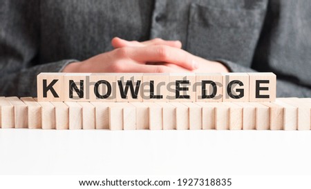 The hand puts a wooden cube with the letter KNOWLEDGE. The word is written on wooden cubes standing on the white surface of the table.