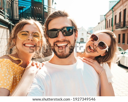 Group of young three stylish friends in the street.Man and two cute girls dressed in casual summer clothes.Smiling models having fun in sunglasses.Women and guy making photo Pov selfie on smartphone