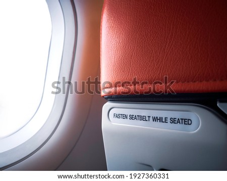 Text message "FASTEN SEAT BELT WHILE SEATED" behind the red airplane seat near the window in cabin of low cost commercial airline for information to passenger for safety emergency with copy space.