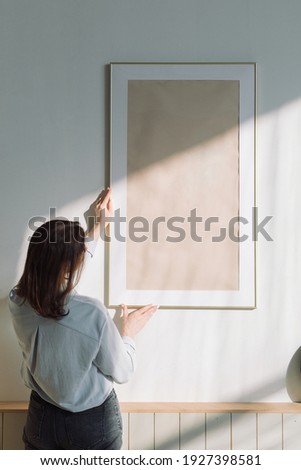 Rear view of a woman hanging a large poster frame on the wall in a modern apartment.