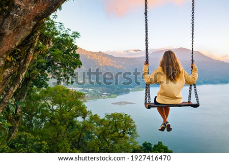 Summer vacation. Young woman sit on tree rope swing on high cliff above tropical lake. Happy girl looking at amazing jungle view. Buyan lake is popular travel destinations in Bali island, Indonesia Royalty-Free Stock Photo #1927416470