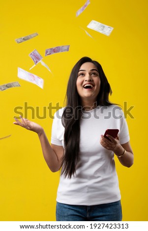 Happy Indian woman using smartphone and flying or raining or falling money, Rupee banknotes on yellow color studio background