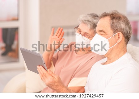 An elderly couple with medical masks sits on the couch at home and communicates with relatives online using a tablet computer. Ways not to get bored during lockdown and develop while sitting at home.