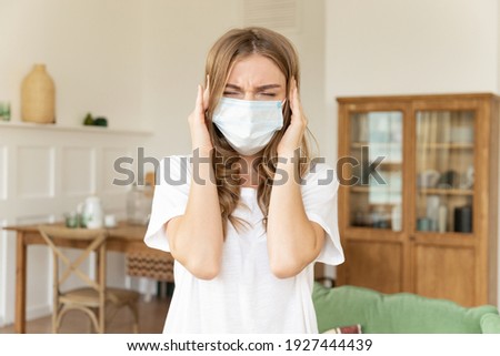 Close up portrait frowning stressed unhappy young woman wearing medical facial mask at home. Worrying lady with covered mouth and nose with closed eyes touching her temples because of headache.