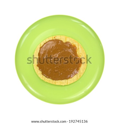 Top view of a corn tortilla with peanut butter on a green plate atop a white background.