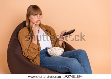 Photo of puzzled charming young lady bite lip switch channel sit chair eat popcorn isolated on pastel beige color background
