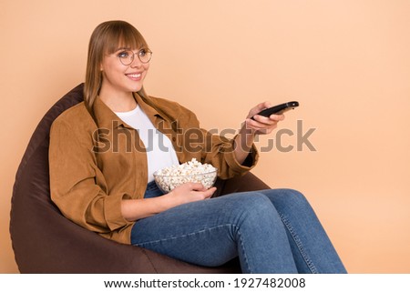 Photo of happy cheerful nice woman hold remote control channel sit bean bag eat pop corn isolated on beige color background