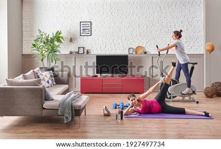 Two girl friends are doing training and sport at home, stretching and cycling concept. Decorative living room, brick wall and television.