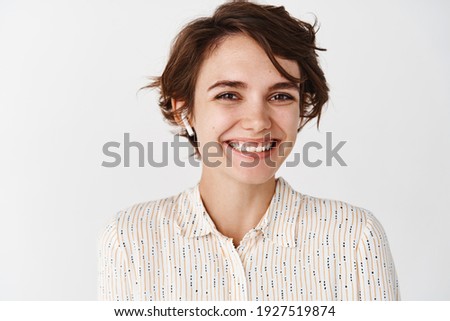 Close-up of beautiful candid woman smiling and looking happy, listening music or podcast in wireless earphones, wearing headphones on white background.