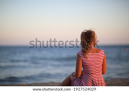 Beituful Woman in red-and-white dress siting by back and looking far away. Sea. Calmness. Romantic. Sunset.