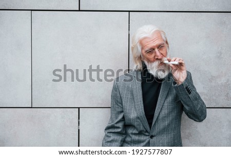 Smoking electronic cigarette. Senior businessman in formal clothes, with grey hair and beard is outdoors.