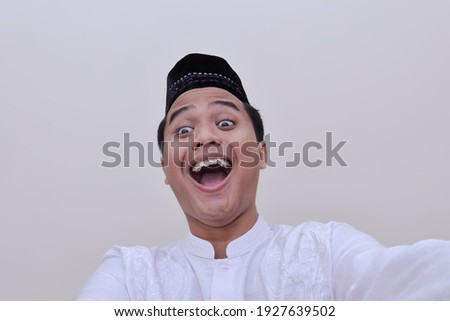 Portrait of religious Asian man in koko shirt or white muslim shirt and black cap taking picture of himself or selfie with eyes bulging. Isolated image on white