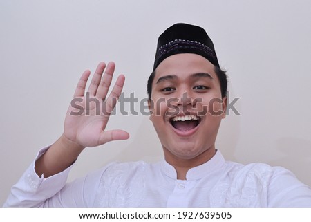 Portrait of religious Asian man in koko shirt or white muslim shirt and black cap taking picture of himself or selfie, saying hi and waving his hand. Isolated image over white background