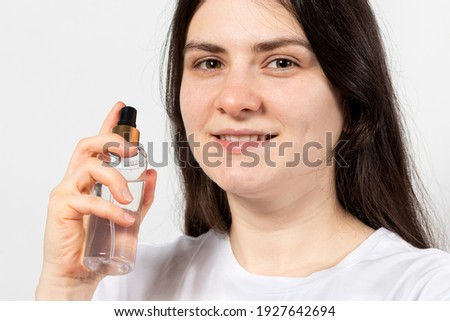 A young brunette woman holds in her hand a bottle with a tonic, hydrolate or aerosol natural cosmetics on a white background with place fo text copy space. Facial and hair care in cosmetology Royalty-Free Stock Photo #1927642694