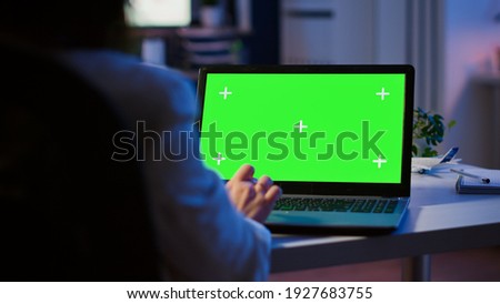 Close up of freelancer typing on laptop with green screen sitting at desk in start up office overworking. Manager watching desktop monitor display with green mockup, chroma key, during night time