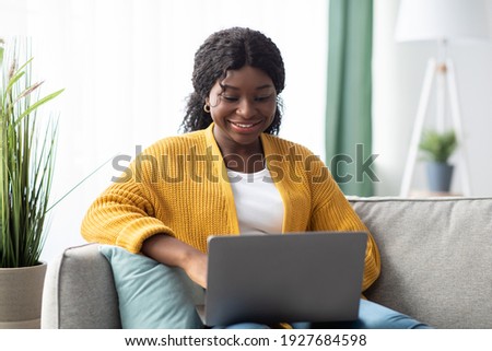 Smiling young black woman in knitted sweater freelancer working from home, sitting on couch, using laptop, copy space. Relaxed african american lady enjoying her weekend, surfing on internet on laptop Royalty-Free Stock Photo #1927684598