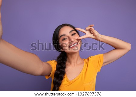 Portrait of smiling young indian woman taking selfie picture, point of view pov shot of happy lady looking at camera making front selfportrait at purple studio, showing v victory sign peace gesture